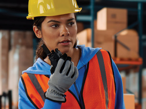 A warehouse worker wearing a yellow hard hat and an orange high-visibility safety vest holding a walkie-talkie.
