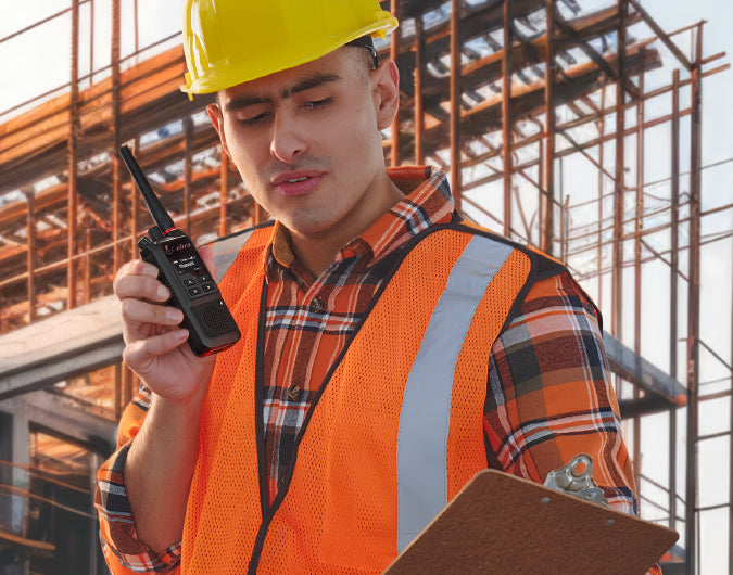 A construction worker wearing a yellow hard hat and an orange safety vest holding a walkie-talkie and a clipboard on a scaffolding site.