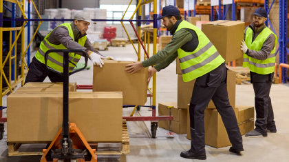 Three warehouse workers in high-visibility yellow vests loading a cardboard box onto a pallet.