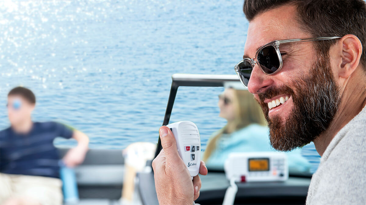Man smiling, wearing sunglasses on a boat using a Cobra BlueBound 1350 marine radio with passengers in the background