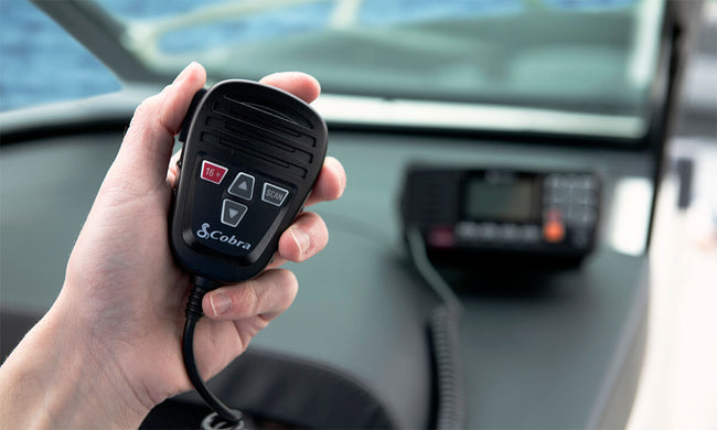 Hand holding the microphone of a Cobra BlueBound 1350 with the fixed mount radio on the boat dashboard in the background