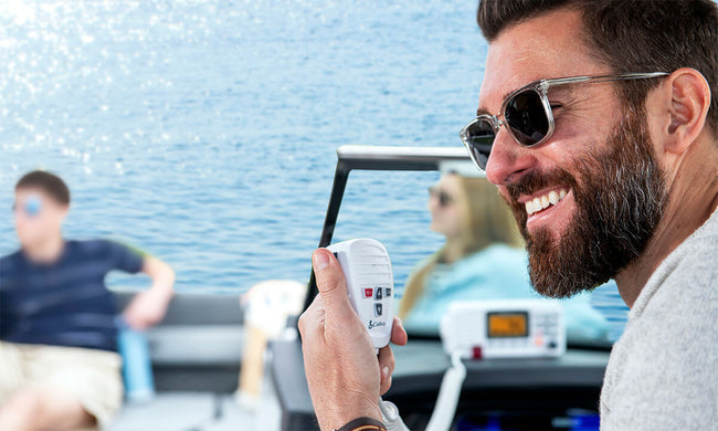 Man wearing sunglasses and smiling in the helm of a boat while holding a white Cobra BlueBound 1350 marine radio device