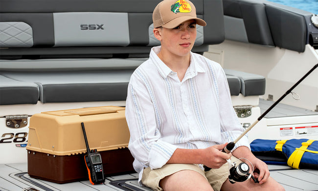 Young man sitting on a boat holding a fishing rod, wearing a Bass Pro Shop cap and a white shirt with a black Cobra BlueBound 350 marine radio