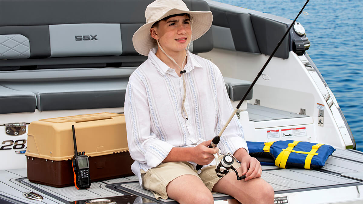 Person sitting on the back of a boat holding a fishing rod, wearing a bucket hat and white shirt with a black Cobra BlueBound 650 handheld marine radio leaning against a brown tacklebox