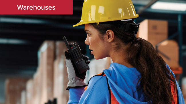 Warehouse staff in yellow hardhat and safety gear uses Cobra PX650 Business Radio to communicate with her team